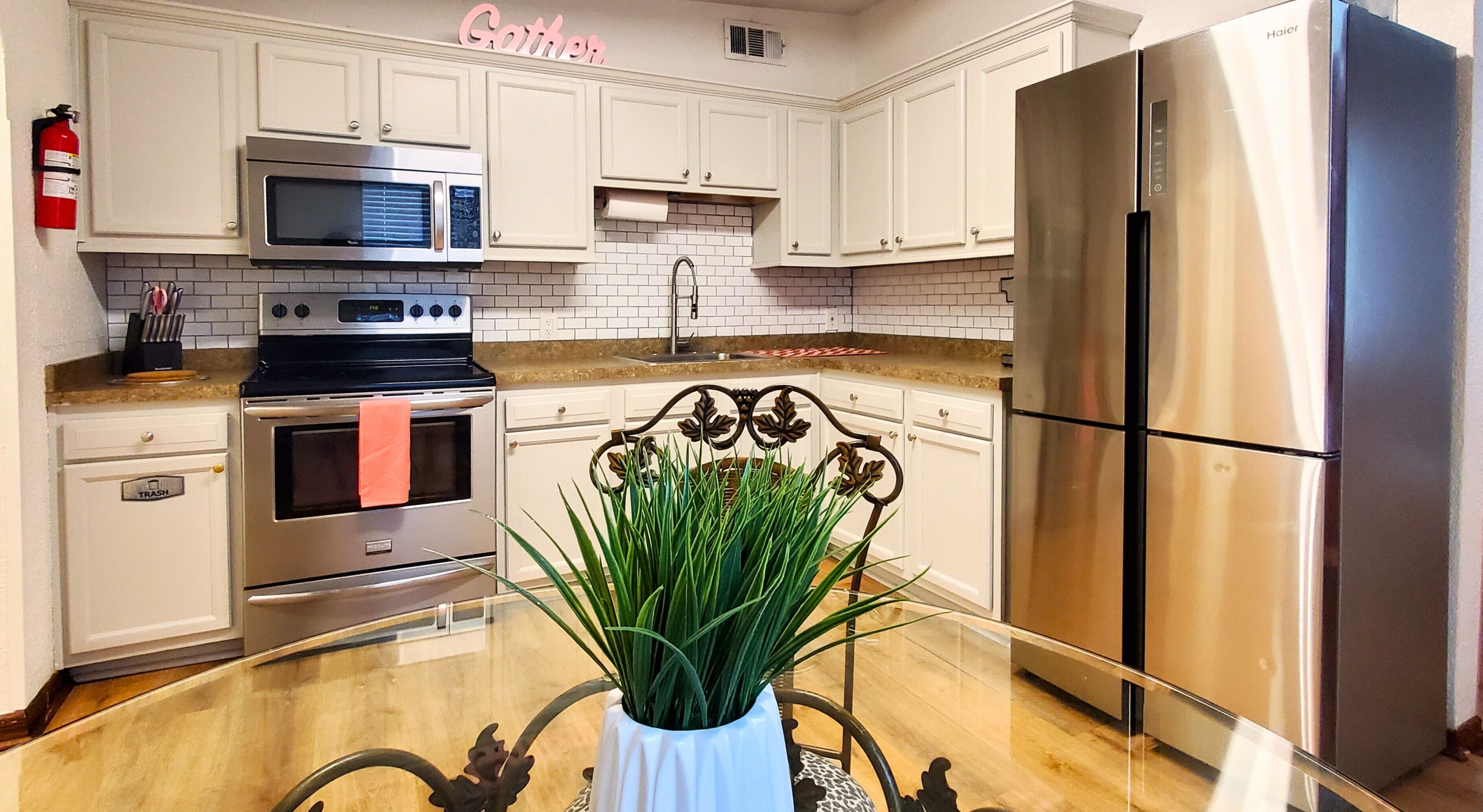 Una cocina moderna con armarios blancos, electrodomésticos de acero inoxidable y una mesa de comedor de vidrio con una planta en maceta.