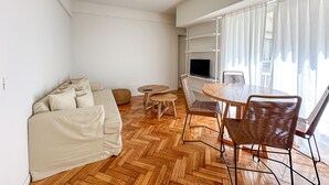 A modern living space with a sofa, a round dining table, and chairs, a television on a shelf, and a large window with curtains. - Modern apartment in Recoleta near Recoleta Mall (Buenos Aires)