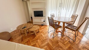 A living room with a wooden herringbone floor, a round wooden table with four chairs, a sofa, a TV on a white shelf, and a window with sheer curtains. - Modern apartment in Recoleta near Recoleta Mall (Buenos Aires)