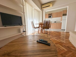 A modern living room with a flat-screen TV, wooden flooring, a dining area with a table and chairs, and a kitchen in the background.