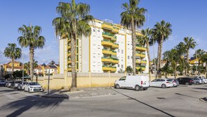 A multi-story building with balconies, parked cars, and palm trees.
