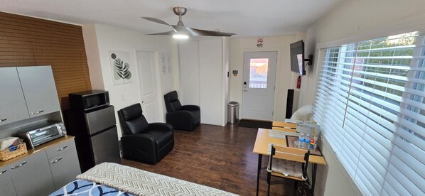 Habitación de hotel compacta con kitchenette, dos sillones de cuero negro, ventilador de techo, televisor montado en la pared y ventana con persianas.