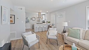 Una sala moderna con un sofá, dos sillones, una mesa de centro y una cocina al fondo.