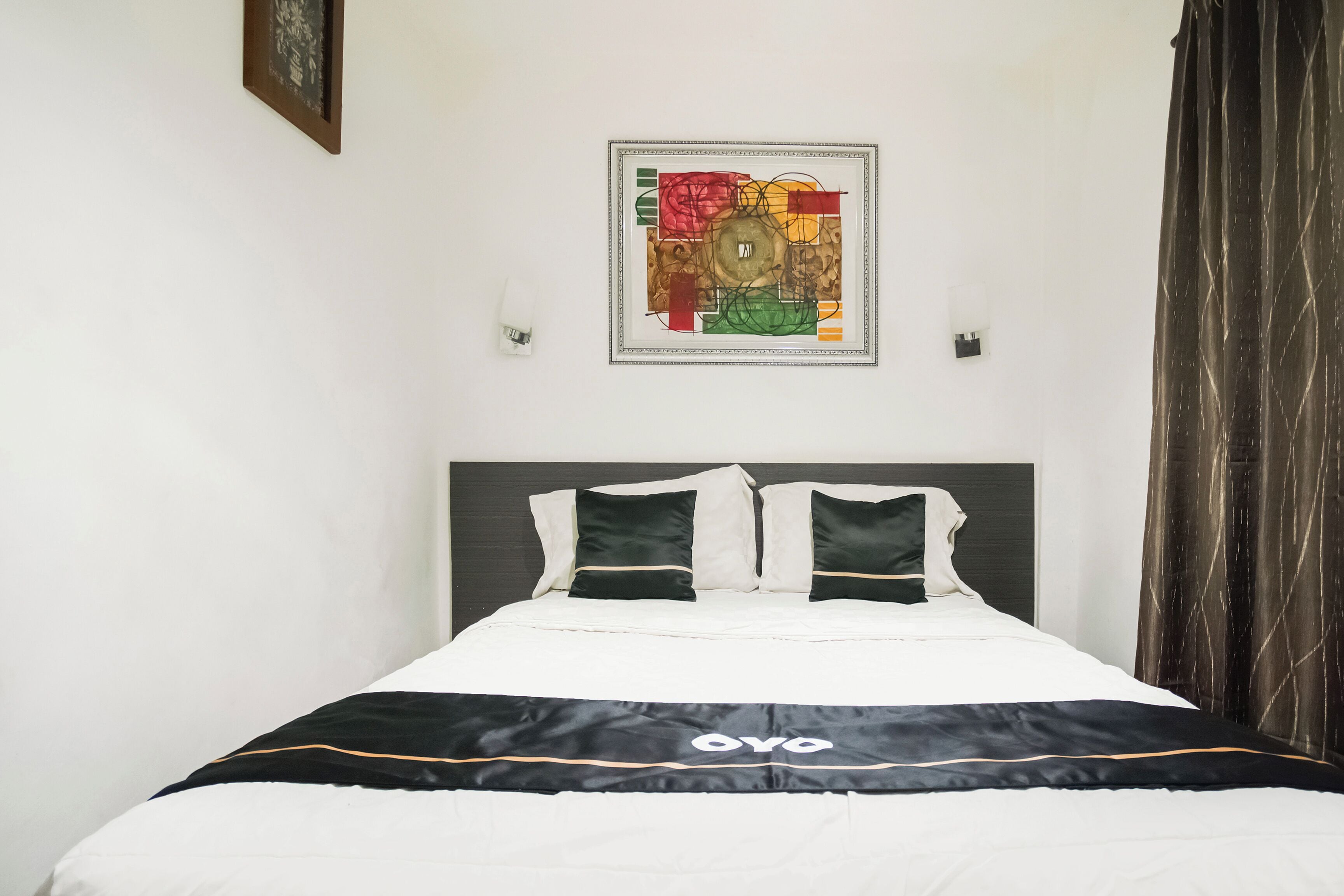 Un lit bien fait, recouvert d’une couvre-lit noir et blanc, avec une œuvre d’art abstraite encadrée au-dessus, et un rideau foncé sur le côté droit.