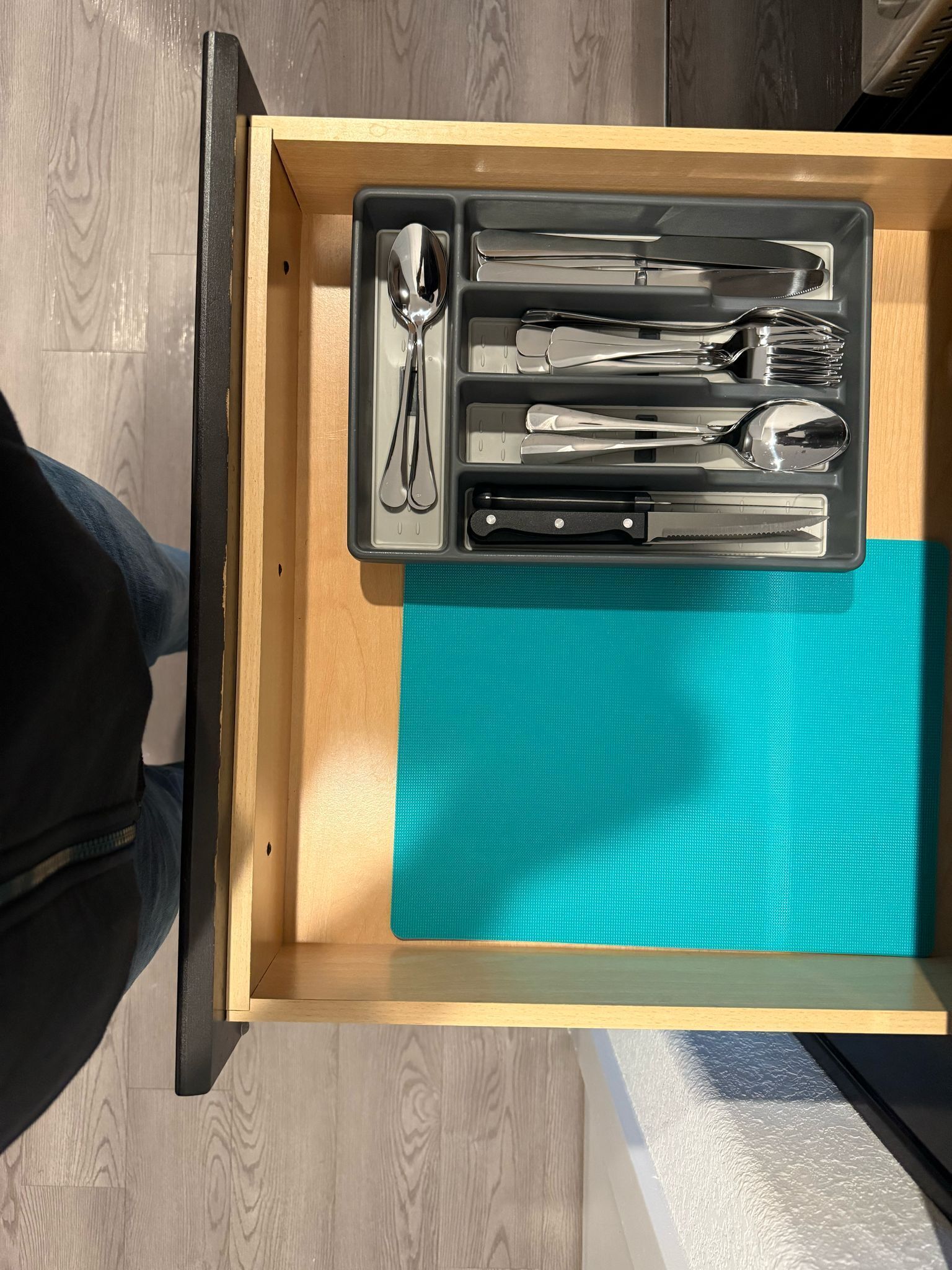 Un cajón con cubiertos, incluyendo cucharas, tenedores y cuchillos, y un tapete color turquesa en su interior.