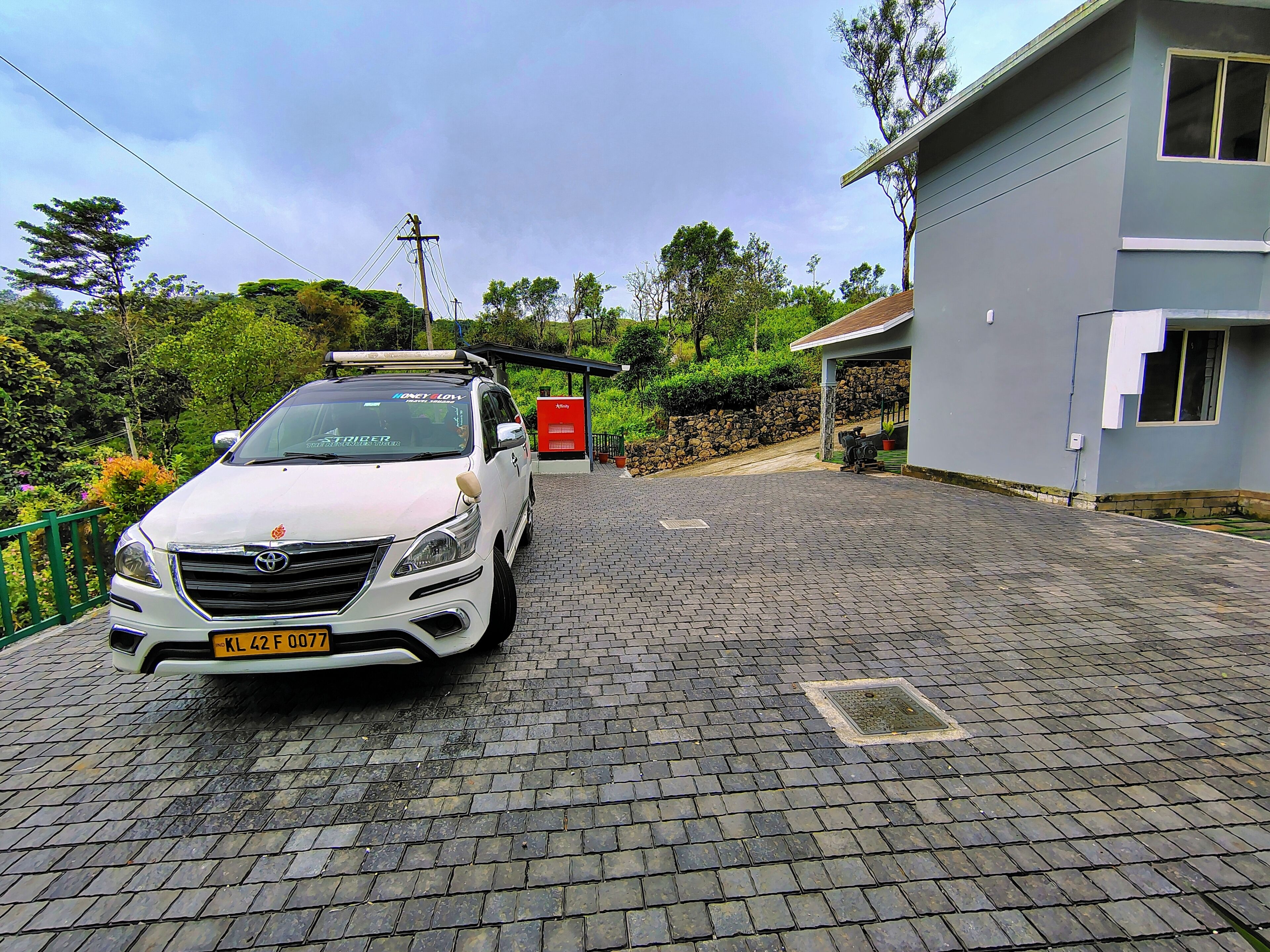 Un SUV blanco estacionado en un camino adoquinado frente a una casa moderna de dos pisos, con un muro de piedra y un cuadro eléctrico rojo.