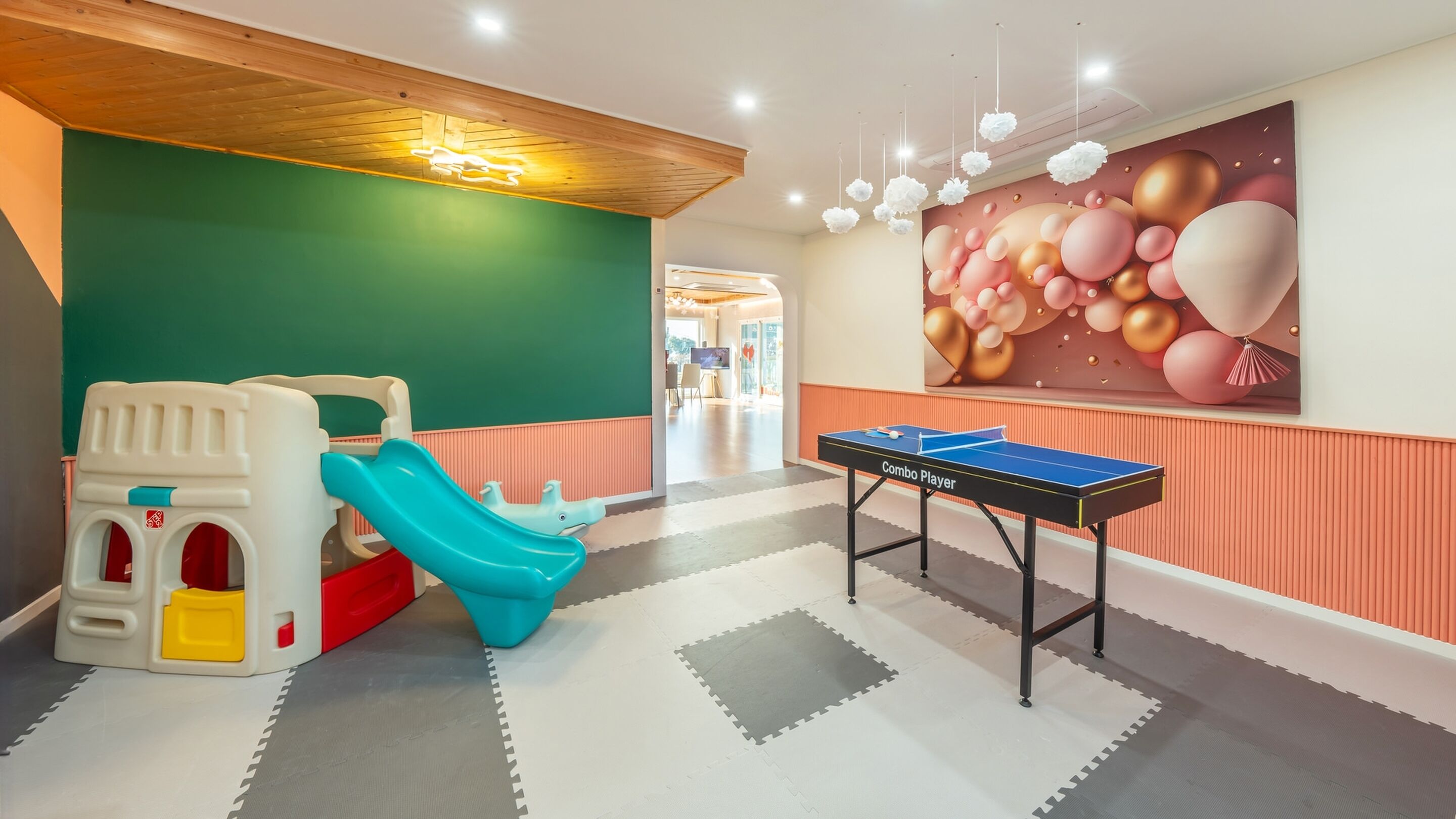 A play area with a slide and a ping pong table in a brightly lit room with a green wall and a balloon mural.