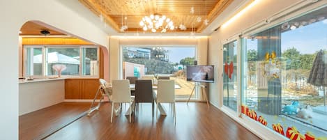 A modern room with a wooden ceiling, a large window with a view of a building and trees, a television, and a dining area with chairs and a table.