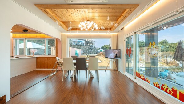 A modern room with a wooden ceiling, a large window with a view of a building and trees, a television, and a dining area with chairs and a table. - Yeosu La Rosa Private Pool Villa Pension (Yeosu)