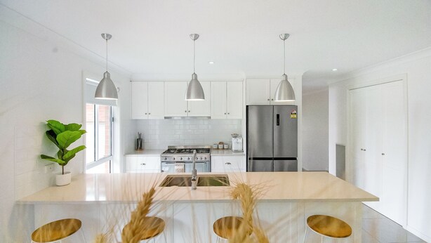 Cocina moderna con gabinetes blancos, electrodomésticos de acero inoxidable, iluminación colgante y una isla central con taburetes.