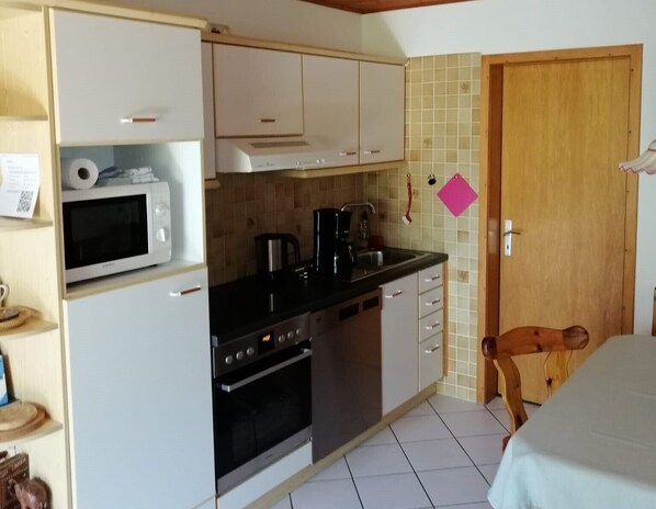 Cocina compacta con microondas, estufa y cafetera. Se ve una puerta de madera y una pequeña mesa con una silla.