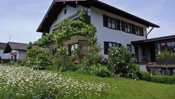 A two-story house with a balcony, surrounded by a well-maintained garden with white flowers and green shrubs.