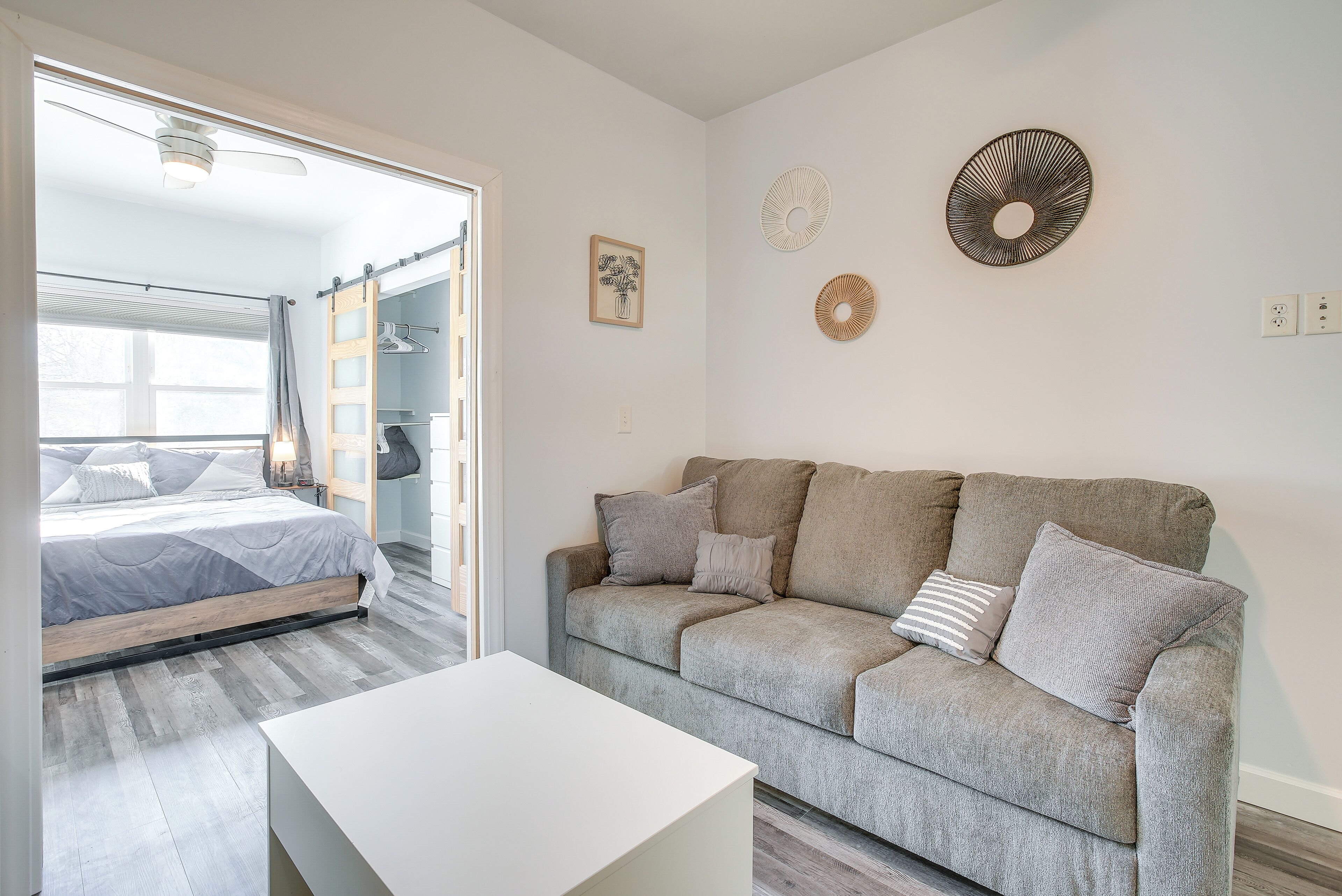 Uma sala de estar moderna com um sofá cinzento, uma mesa de centro branca e uma cama com roupa de cama cinza. Há decorações nas paredes e um ventilador de teto.