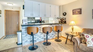 Eine moderne Küche mit weißen Schränken, einer Arbeitsplatte und einer Bar mit Hockern. Im Wohnbereich befindet sich eine Couch und ein kleiner Tisch mit einer Lampe.