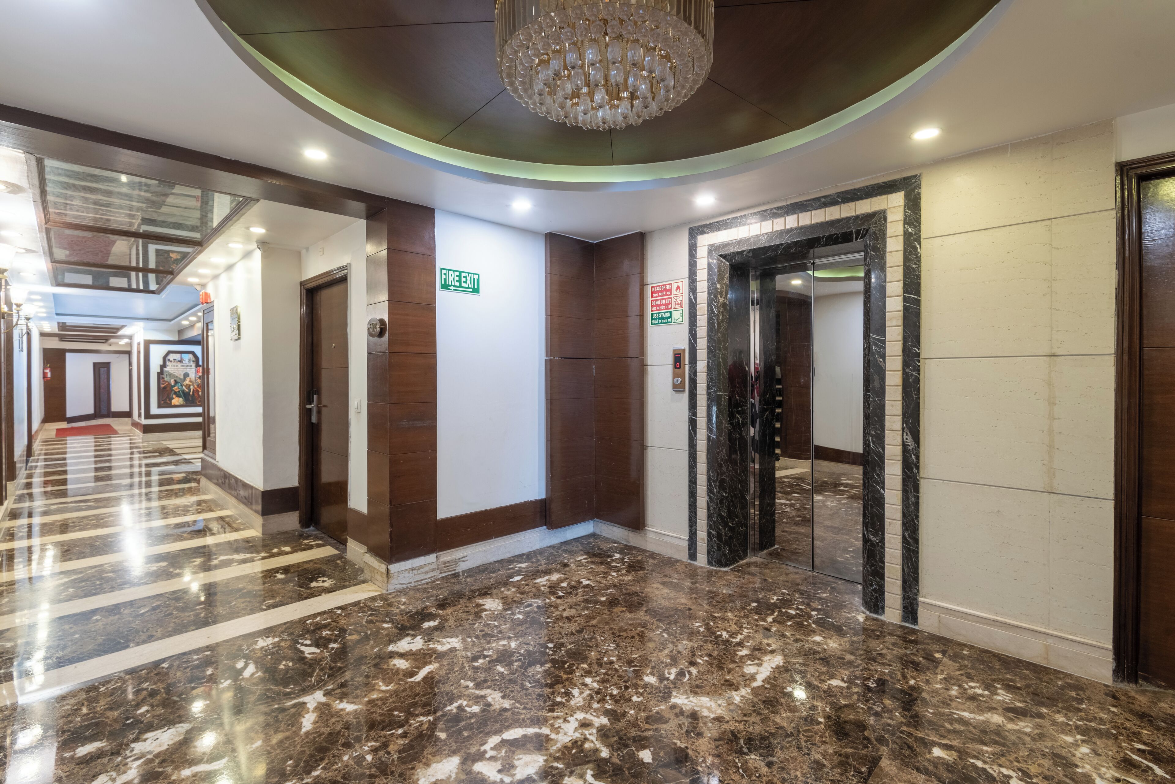 Un hall de hotel con piso de mármol, una araña de cristal, un letrero de salida de incendios y una puerta de ascensor espejada.