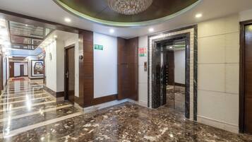 Un hall de hotel con piso de mármol, una araña de cristal, un letrero de salida de incendios y una puerta de ascensor espejada.
