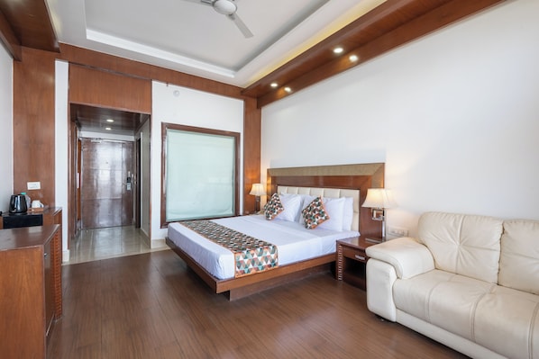 A hotel room with a wooden floor, a bed with a patterned bedspread, a white sofa, a nightstand with a lamp, and a ceiling fan.