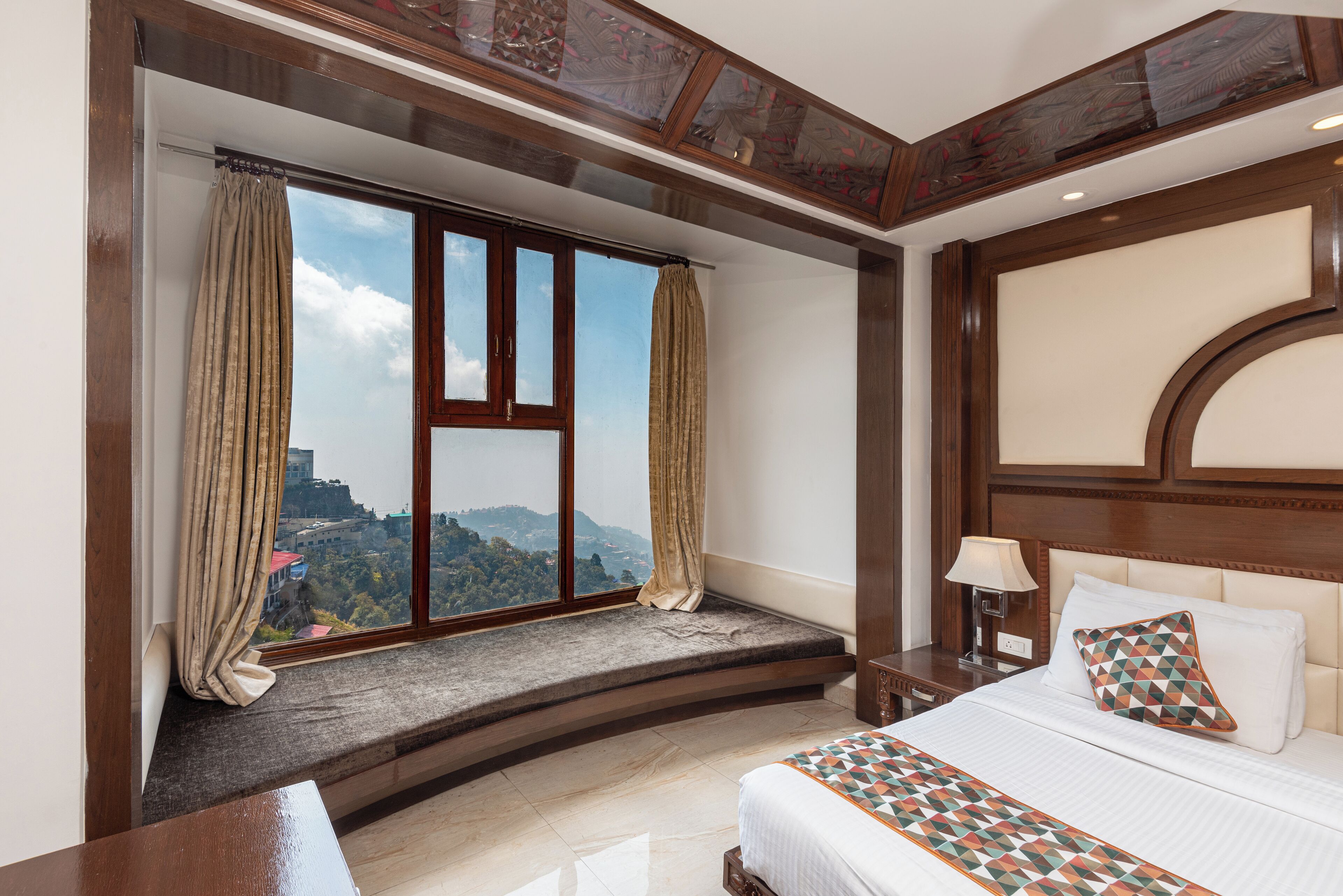A bedroom with a large window, a bed with patterned bedding, a nightstand, and a view of a mountainous landscape.