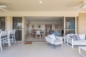 A modern living area with a dining table, sofas, and a sliding glass door leading to a kitchen. - 20 Taine - Oceanstays - Pet-Friendly Waterfront Escape with Fireplace (Yamba)