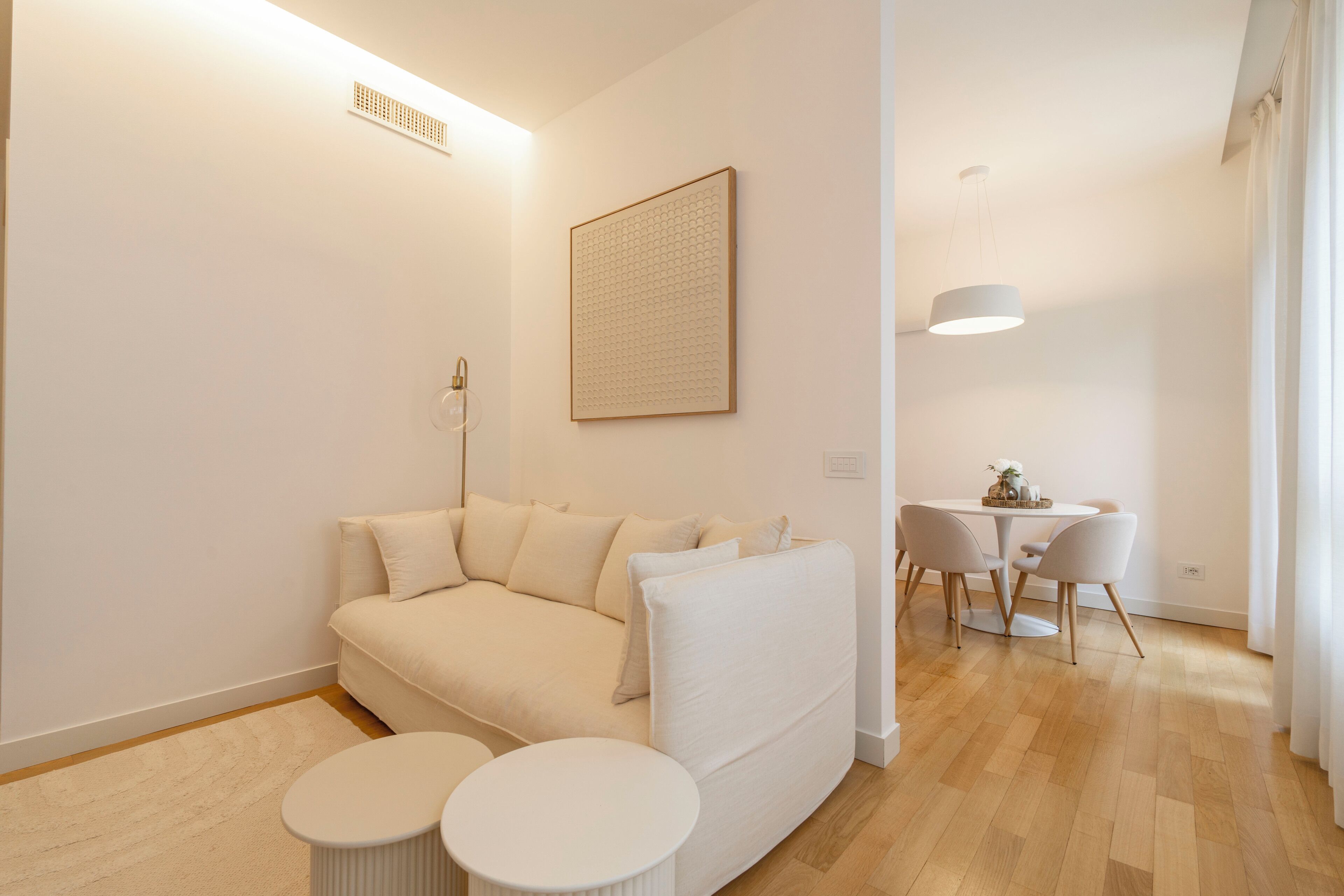 A modern living room with a sofa, two round tables, a dining table with chairs, a pendant light, and a framed artwork on the wall.