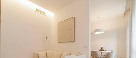 A modern living room with a sofa, two round tables, a dining table with chairs, a pendant light, and a framed artwork on the wall.
