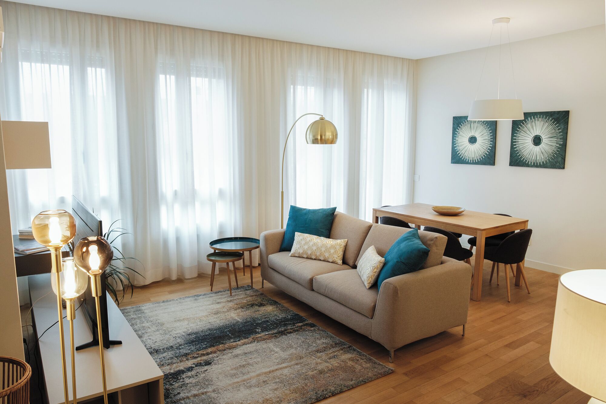 A modern living room with a sofa, coffee table, and dining table. There are two lamps, a rug, and artwork on the wall.