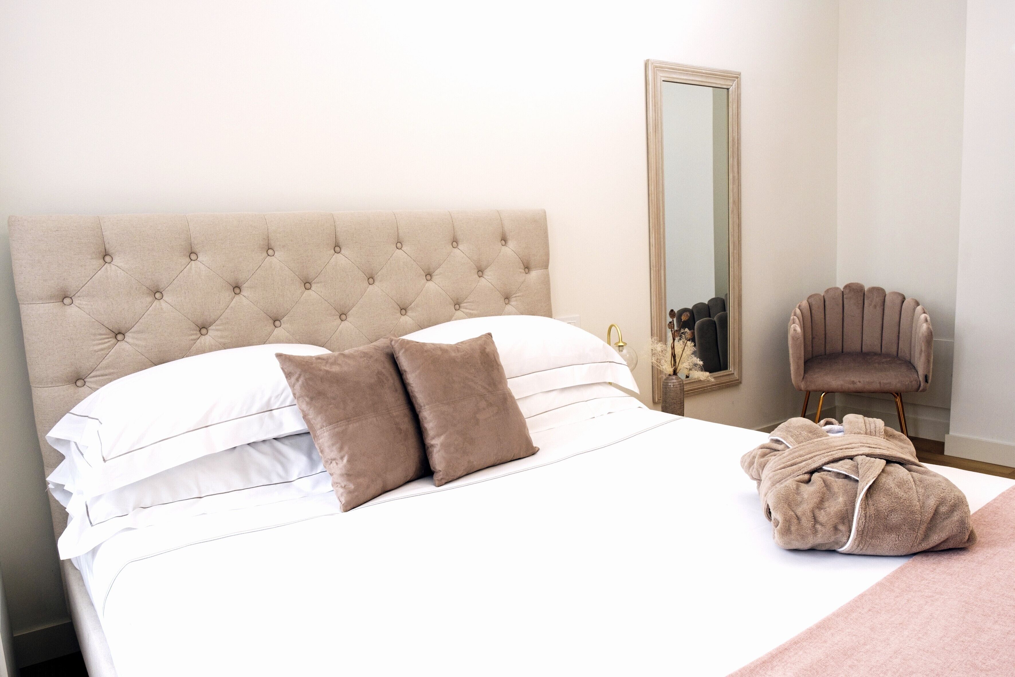 Un lit bien fait, avec une tête de lit capitonnée, un miroir et une chaise, dans une chambre d’hôtel.