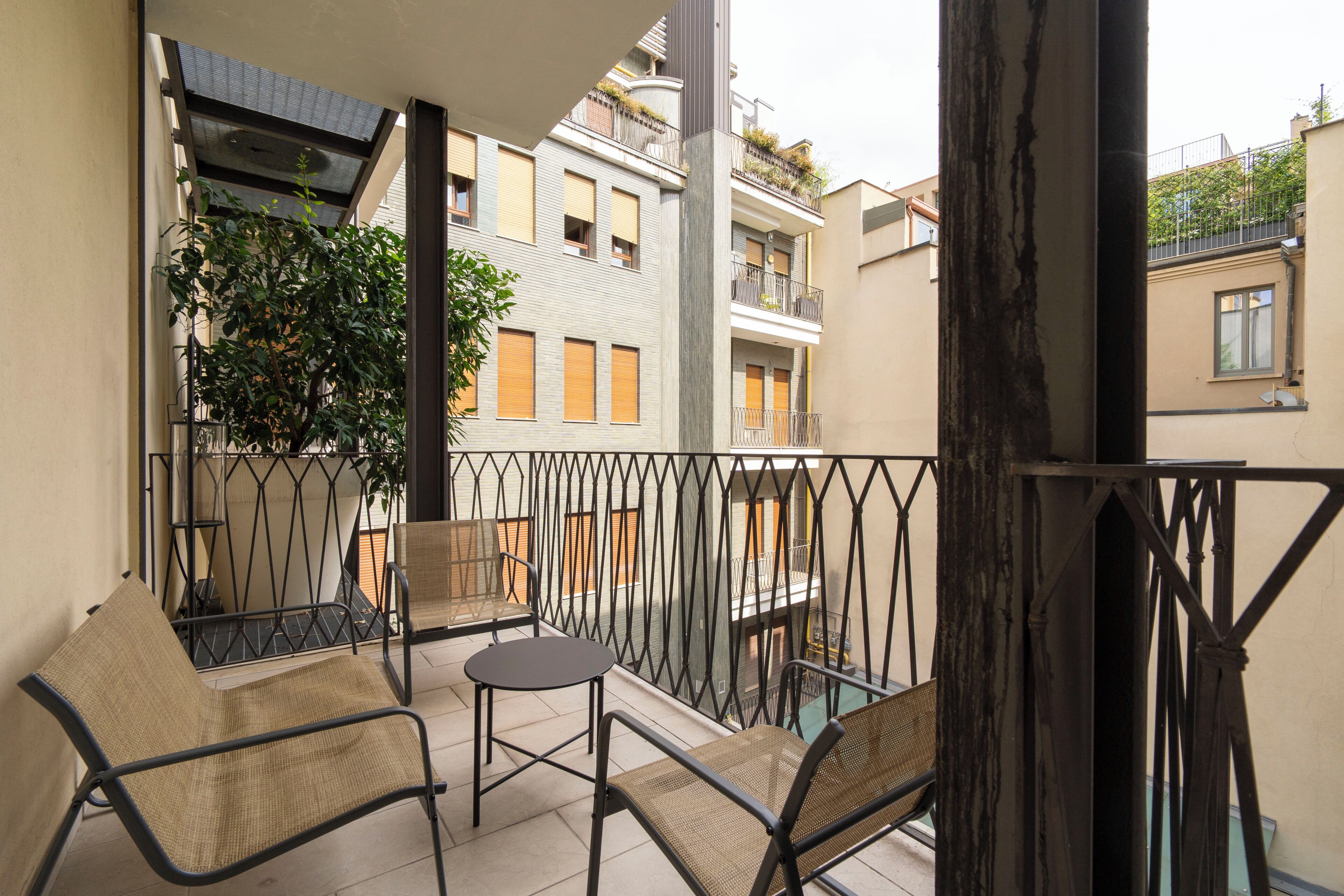 Een balkon met twee stoelen en een tafeltje, met uitzicht op een woonwijk met gebouwen en een boom.
