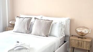 Ein ordentlich bezogenes Bett mit weißen Leinen und mehreren Kissen, ein Nachttisch aus Holz mit einer Lampe und ein Parkettboden.