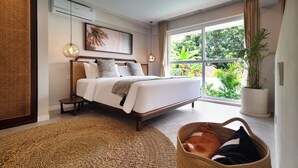 A modern bedroom with a large bed, a small bedside table, a potted plant, and a view of greenery outside. - A.K.A Tropicana Bingin (Pecatu)