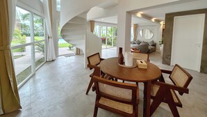A spacious living area with a spiral staircase, a round wooden table with chairs, and large windows offering a view of a garden. - A.K.A Tropicana Bingin (Pecatu)