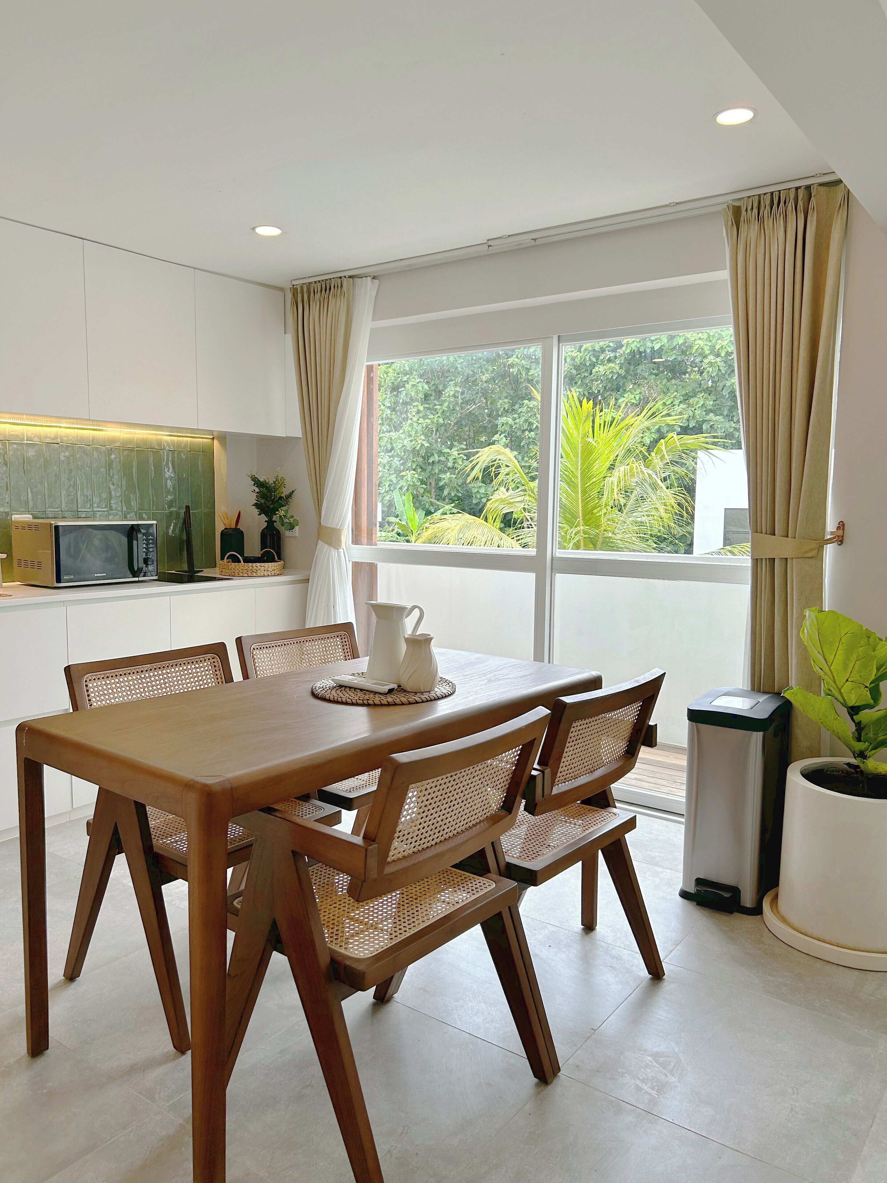 A modern kitchen with white cabinetry, a wooden dining table with wicker chairs, and a view of greenery outside.