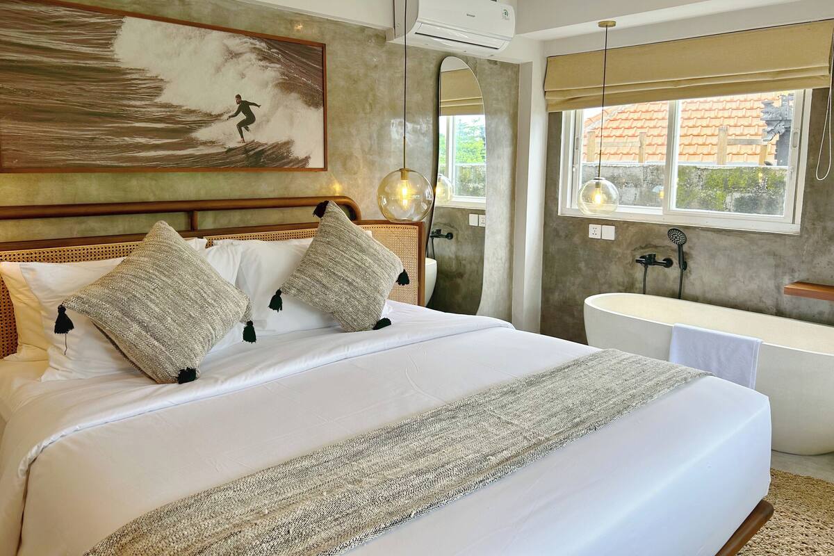 Een slaapkamer met een groot bed, een schilderij, een badkamer met een bad en een raam met uitzicht op gebouwen.
