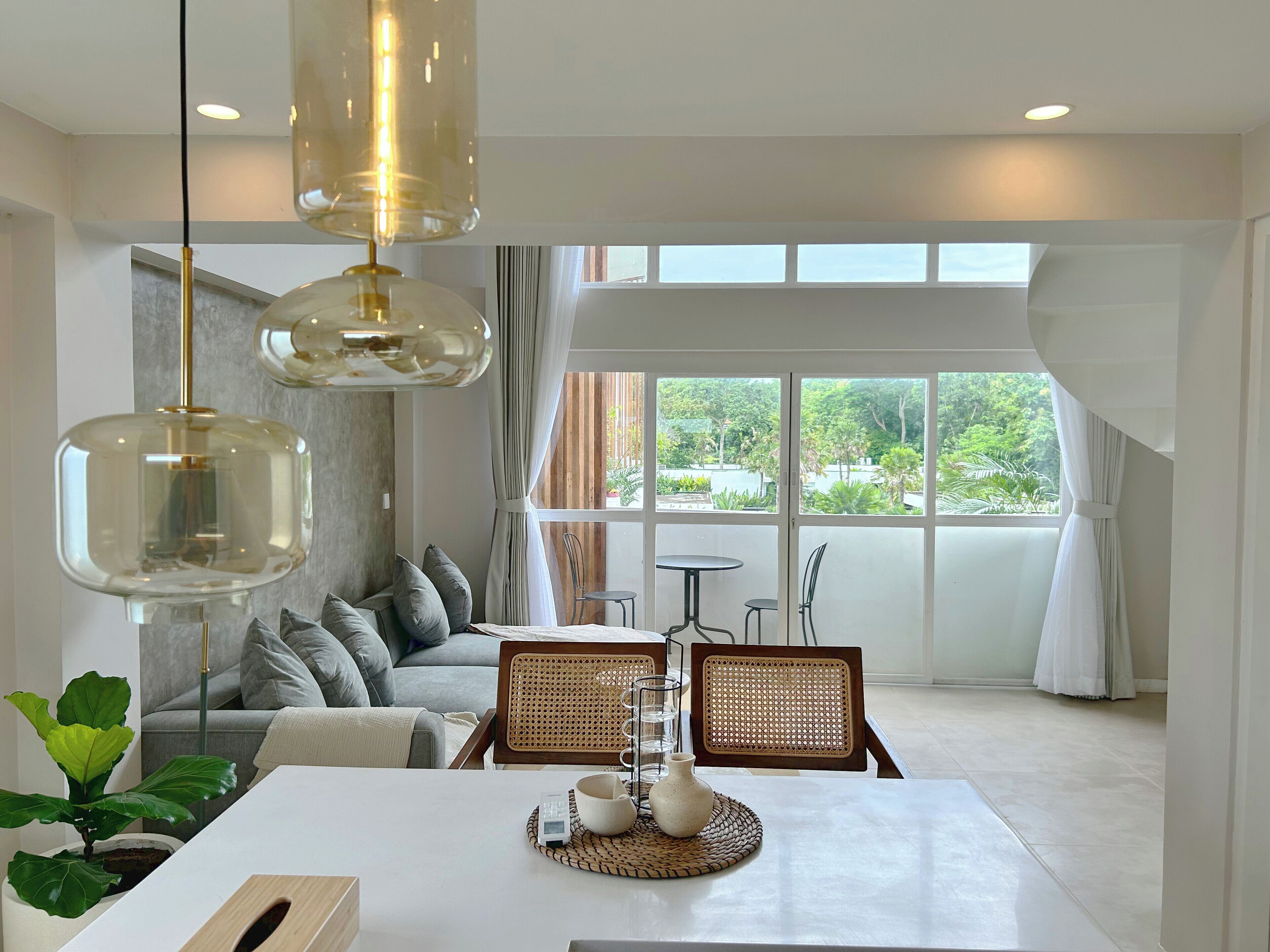 A modern living room with a sofa, dining table, and chairs. There are large windows with curtains, a hanging light fixture, and a potted plant.