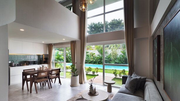 A modern living room with a sofa, dining table, and chairs, a kitchen area, and a large window with a view of a pool and greenery. - A.K.A Tropicana Bingin (Pecatu)
