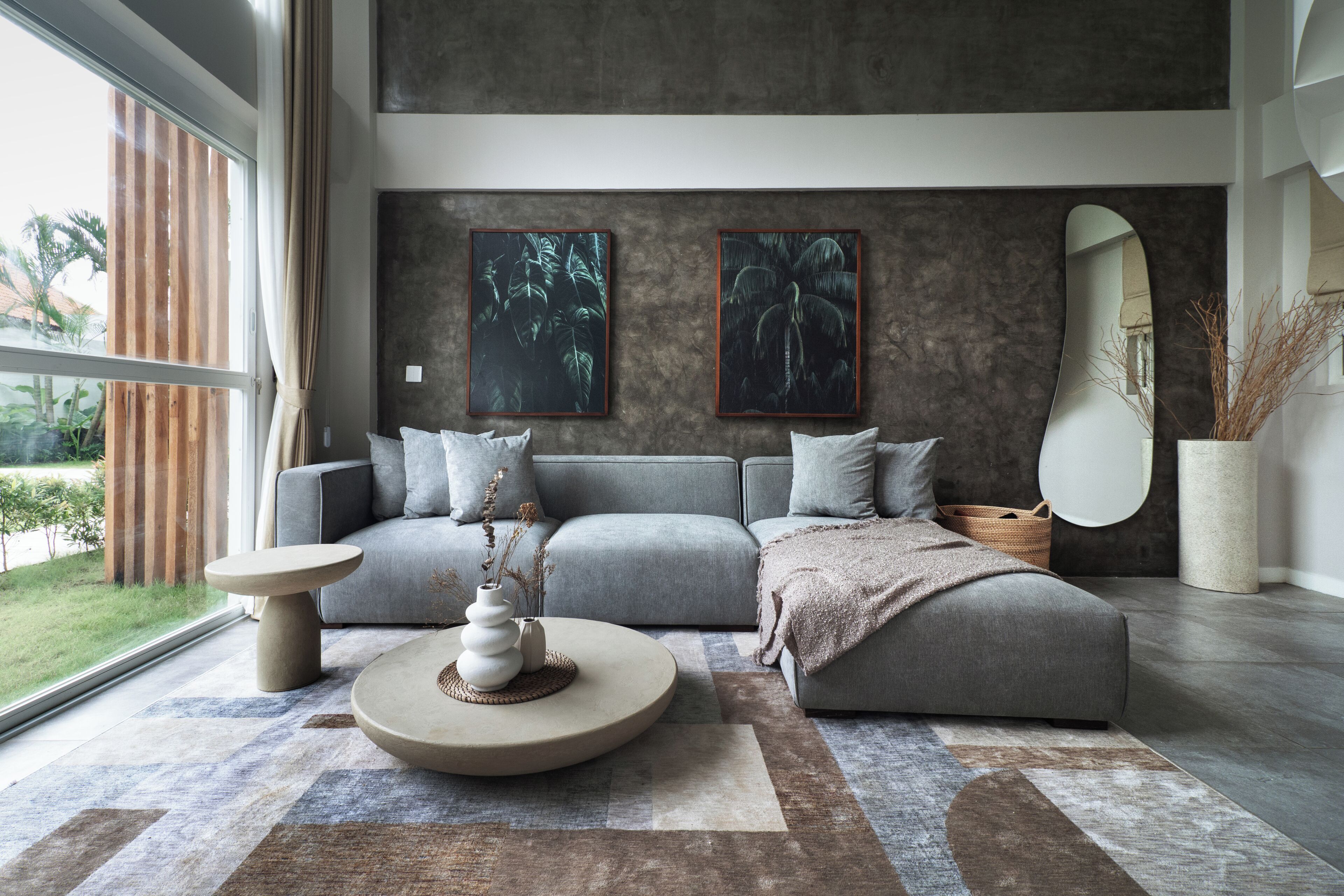 Ein modernes Wohnzimmer mit einem grauen Sofa, zwei Wandgemälden, einem großen Spiegel und einem runden Couchtisch mit einer Vase.