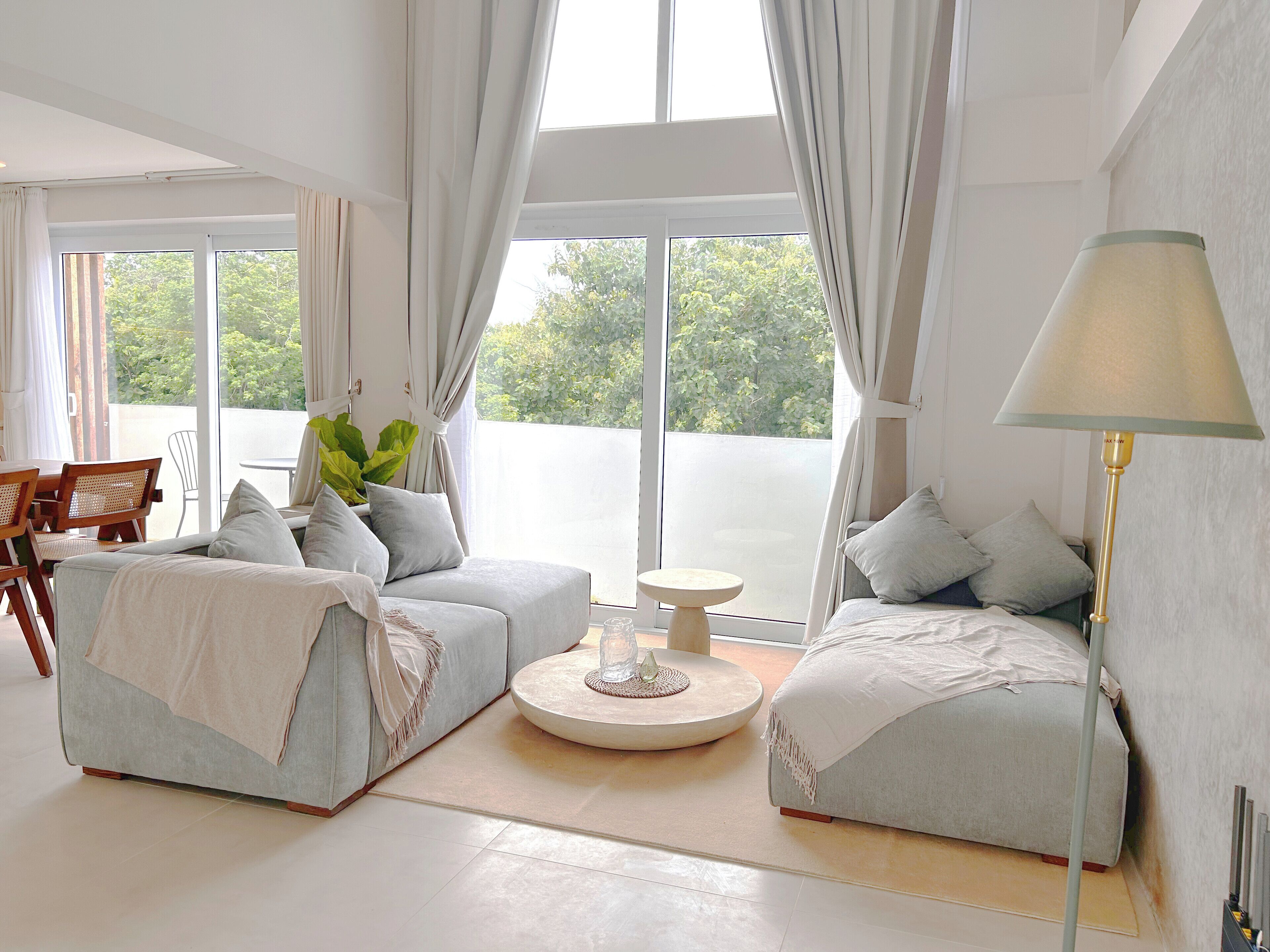 A modern living room with two sofas, a round coffee table, and a lamp. Large windows provide a view of greenery outside.