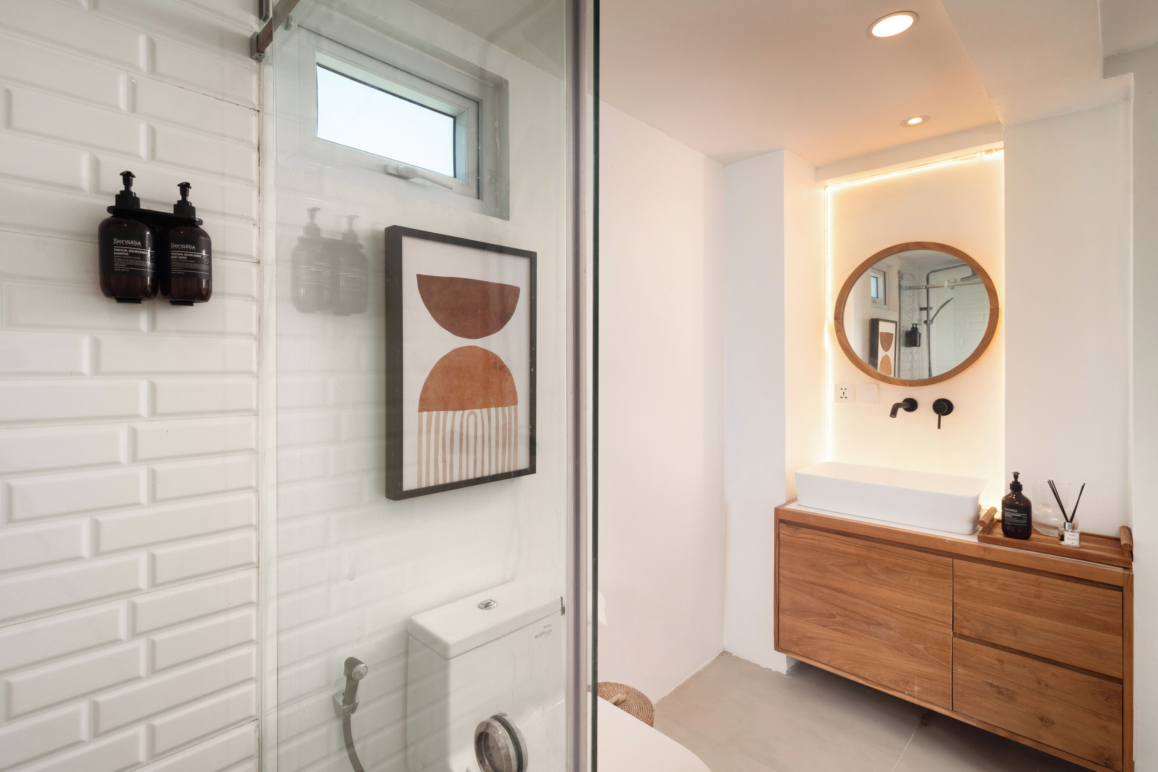 A bathroom with a white brick wall, a glass shower enclosure, a wooden vanity, and a round mirror with a wooden frame.