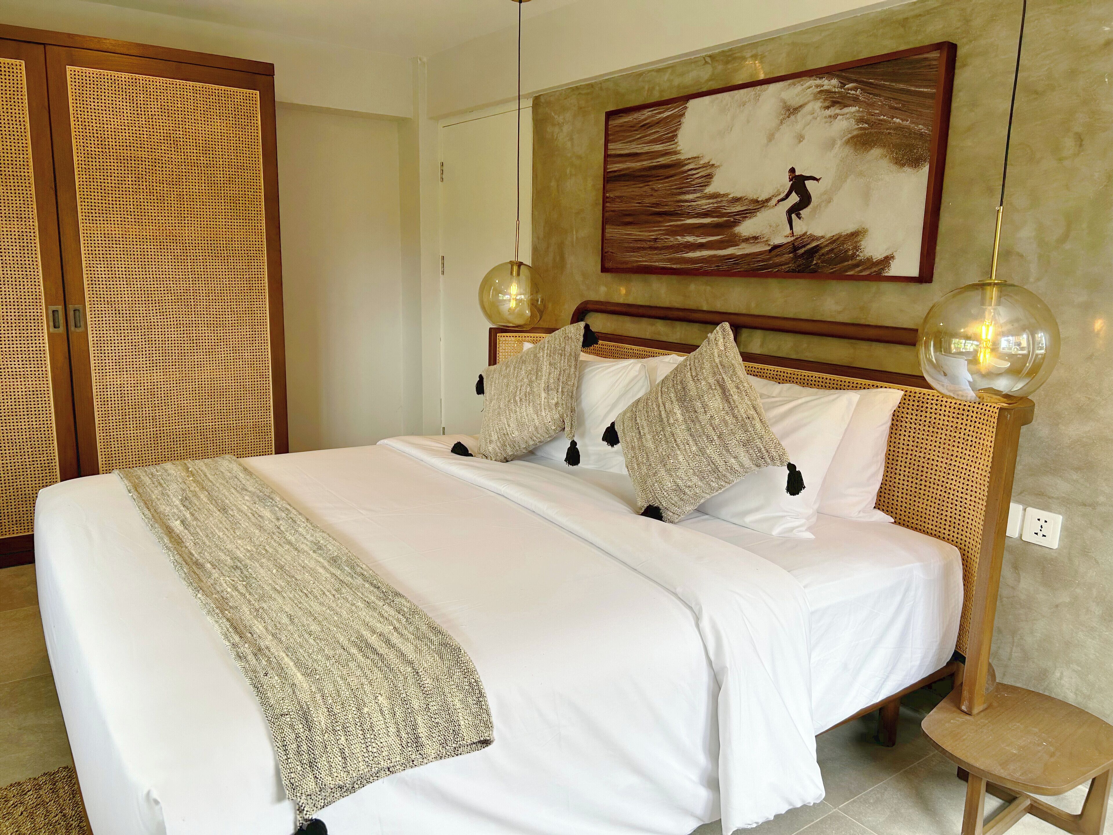 A neatly made bed with white linens and decorative pillows, a wooden headboard, and a painting of a surfer on a wave above it.