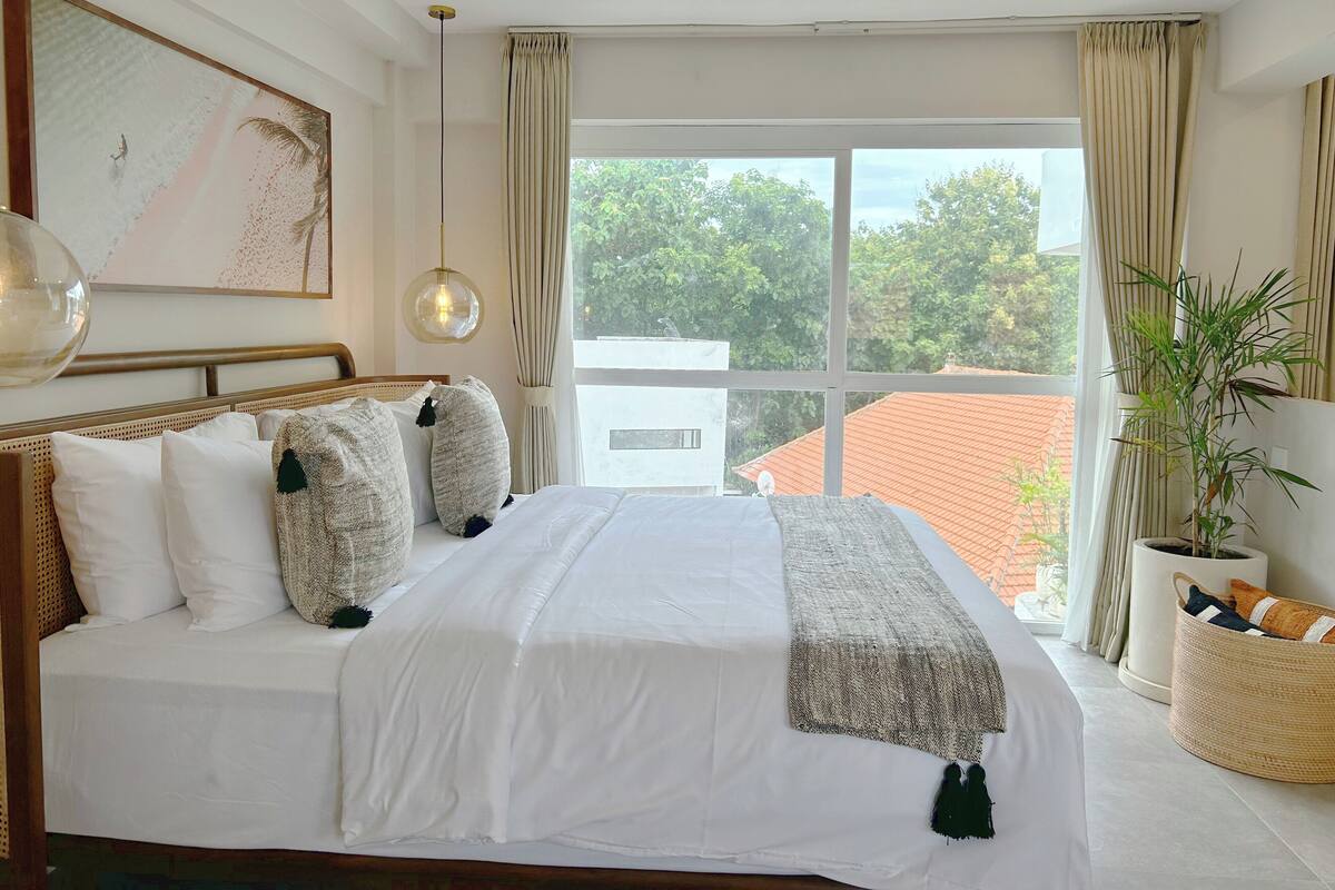Een hotelkamer met een groot bed, een hanglamp, een raam met gordijnen en uitzicht naar buiten.