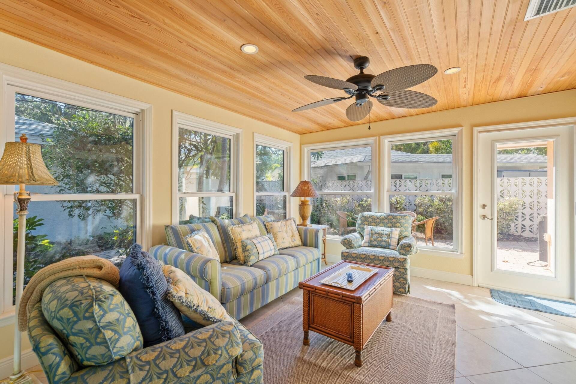 A sunlit room with a wooden ceiling, large windows, a striped sofa, a patterned armchair, and a ceiling fan.
