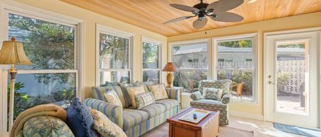 A sunlit room with a wooden ceiling, large windows, a striped sofa, a patterned armchair, and a ceiling fan.