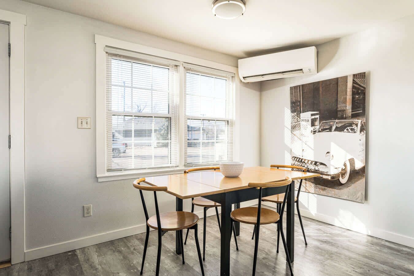 A dining area with a wooden table and four chairs, large windows with blinds, and a vintage car poster on the wall.