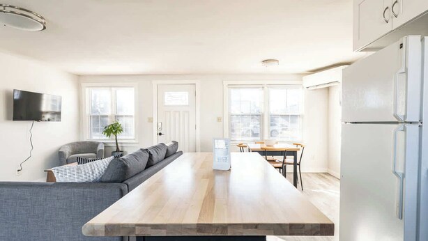 A modern kitchen with a central island, a dining area with a table and chairs, a grey sofa, and a flat-screen TV mounted on the wall. - #StayinMyDistrict Depot Bench Near Downtown BSU (Boise)