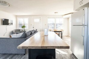Een moderne keuken met een centraal eiland, een eethoek met een tafel en stoelen, een grijze bank en een flatscreen televisie aan de muur.