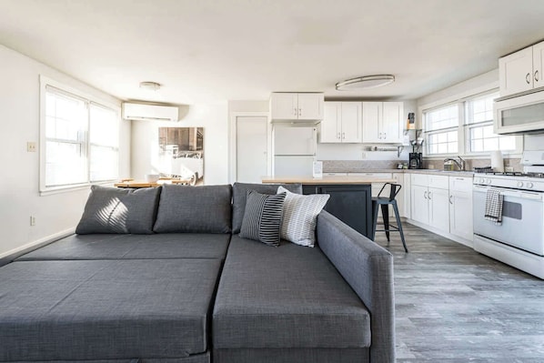 Eine moderne Küche mit weißen Schränken, einem grauen Sofa mit Kissen und einem großen Fenster für natürliches Licht.