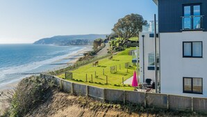 Un bâtiment situé sur la côte, offrant une vue sur la mer, une zone herbeuse avec un banc et une clôture en bois.