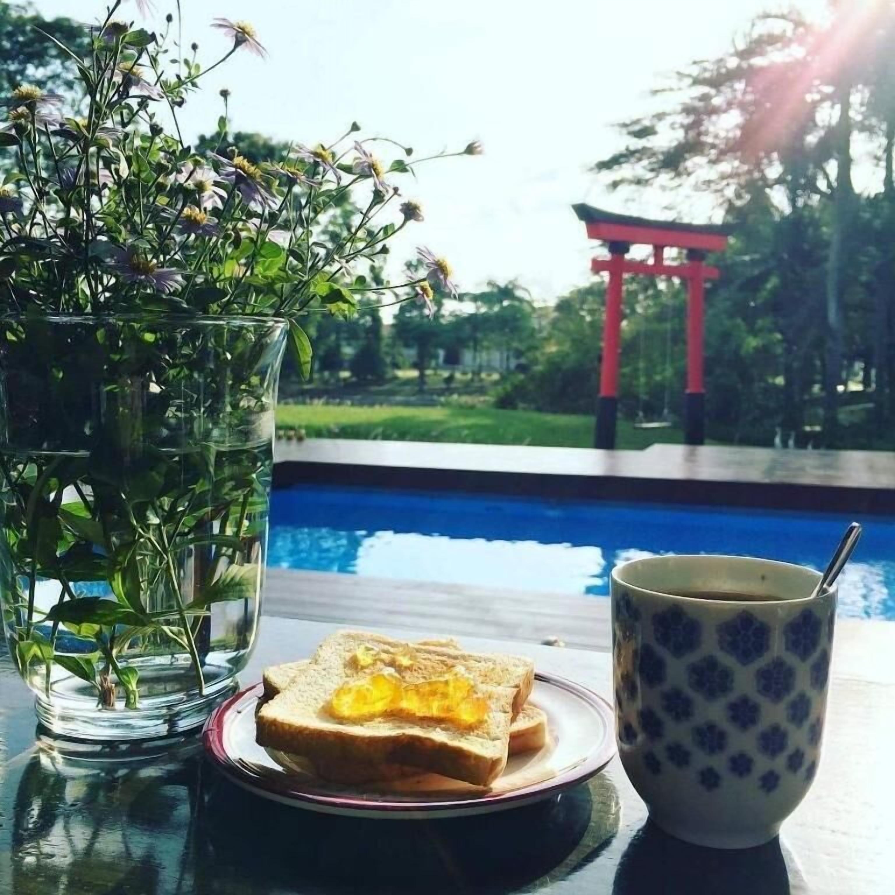 Eine Glasvase mit Blumen, ein Teller mit Toast und eine Tasse auf einem Tisch, neben einem Pool mit einem roten Torii-Tor im Hintergrund.