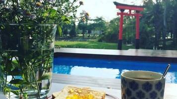 Eine Glasvase mit Blumen, ein Teller mit Toast und eine Tasse auf einem Tisch, neben einem Pool mit einem roten Torii-Tor im Hintergrund.