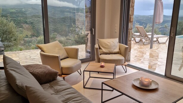 Een moderne woonkamer met een grote bank, twee fauteuils, een salontafel en uitzicht op een bergachtig landschap door grote ramen.